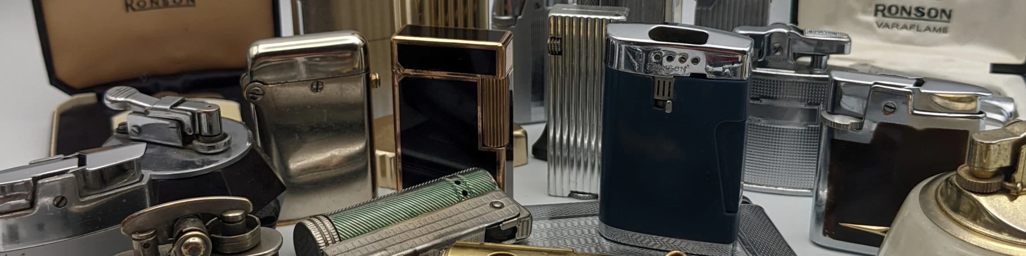 Part of the collection: Ronson, Colibri and Dunhill lighters in chrome, brass and enamel, with original Ronson Varaflame packaging behind.